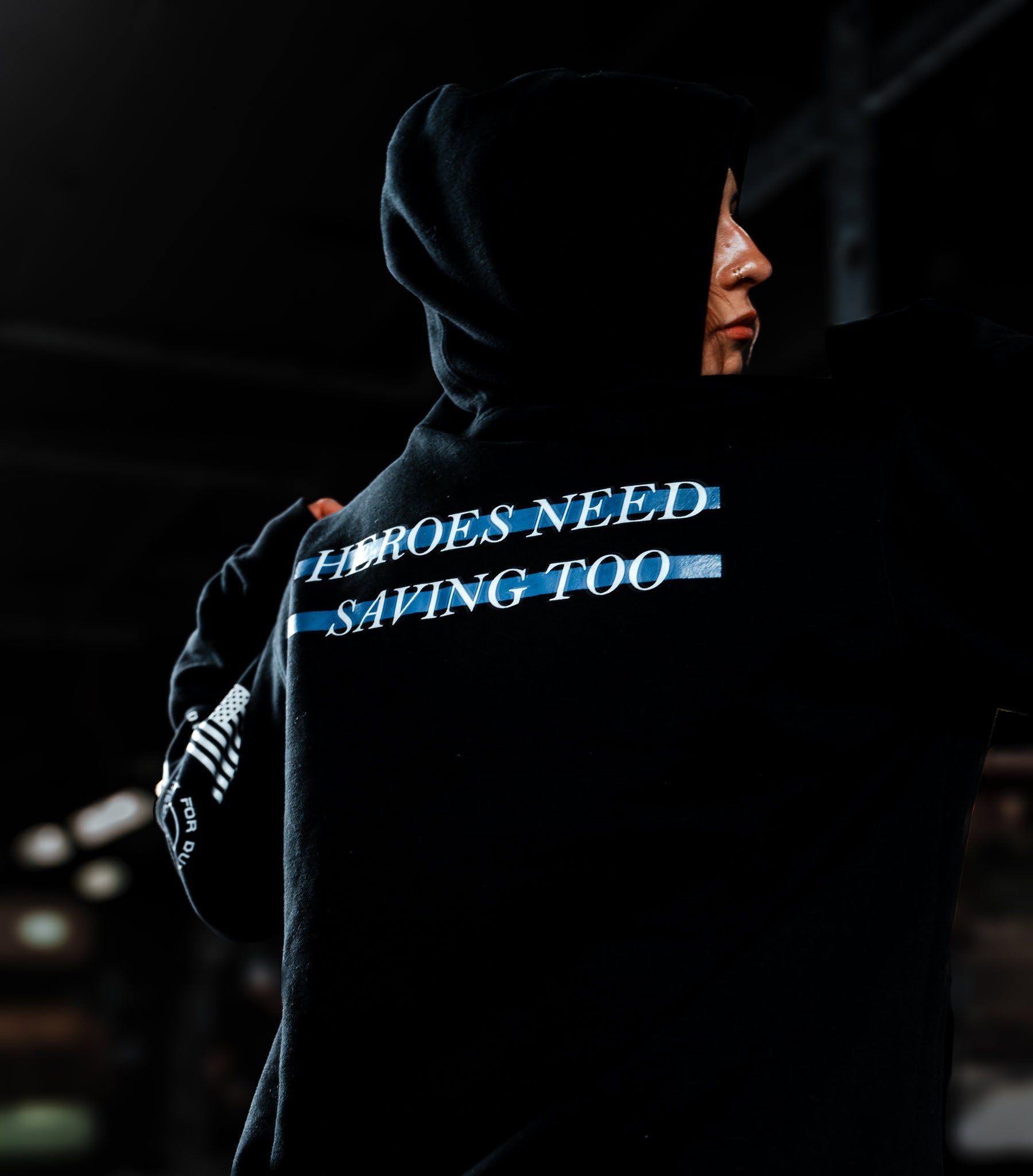Person wearing a hoodie with text 'Heroes Need Saving Too' in a dark setting.