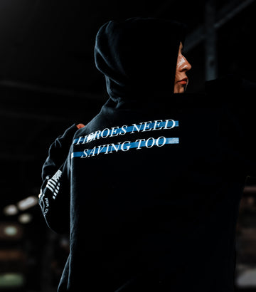 Person wearing a hoodie with text 'Heroes Need Saving Too' in a dark setting.