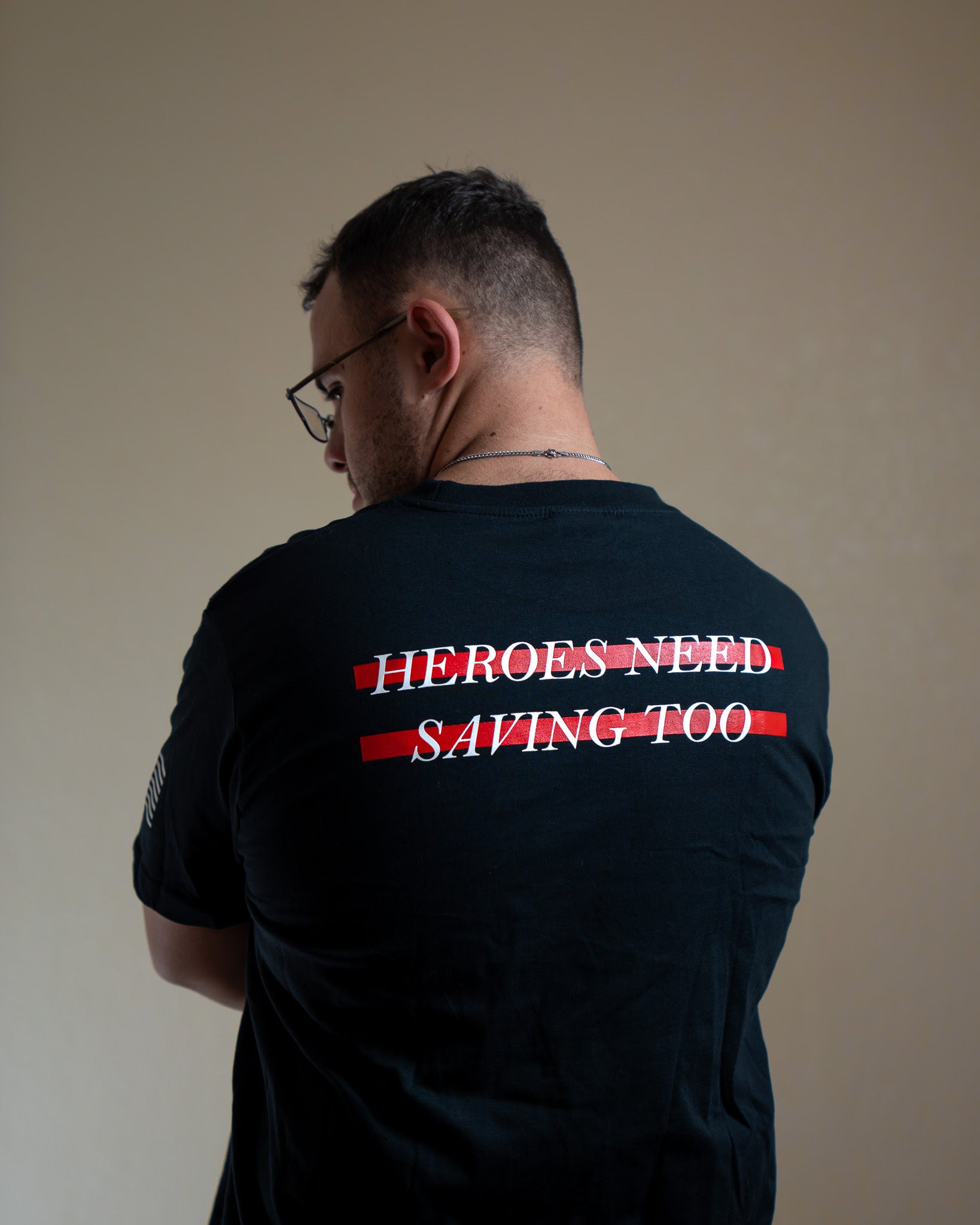Man wearing a black t-shirt with 'HEROES NEED SAVING TOO' text on the back against a plain background