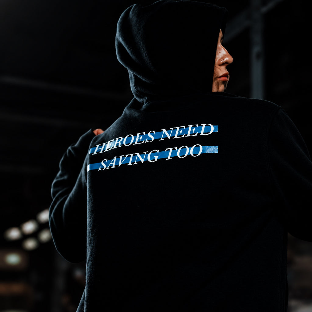 A person wearing a black hoodie, shown from the back, with the text 'HEROES NEED SAVING TOO' printed in white across two blue stripes on the upper back. #black