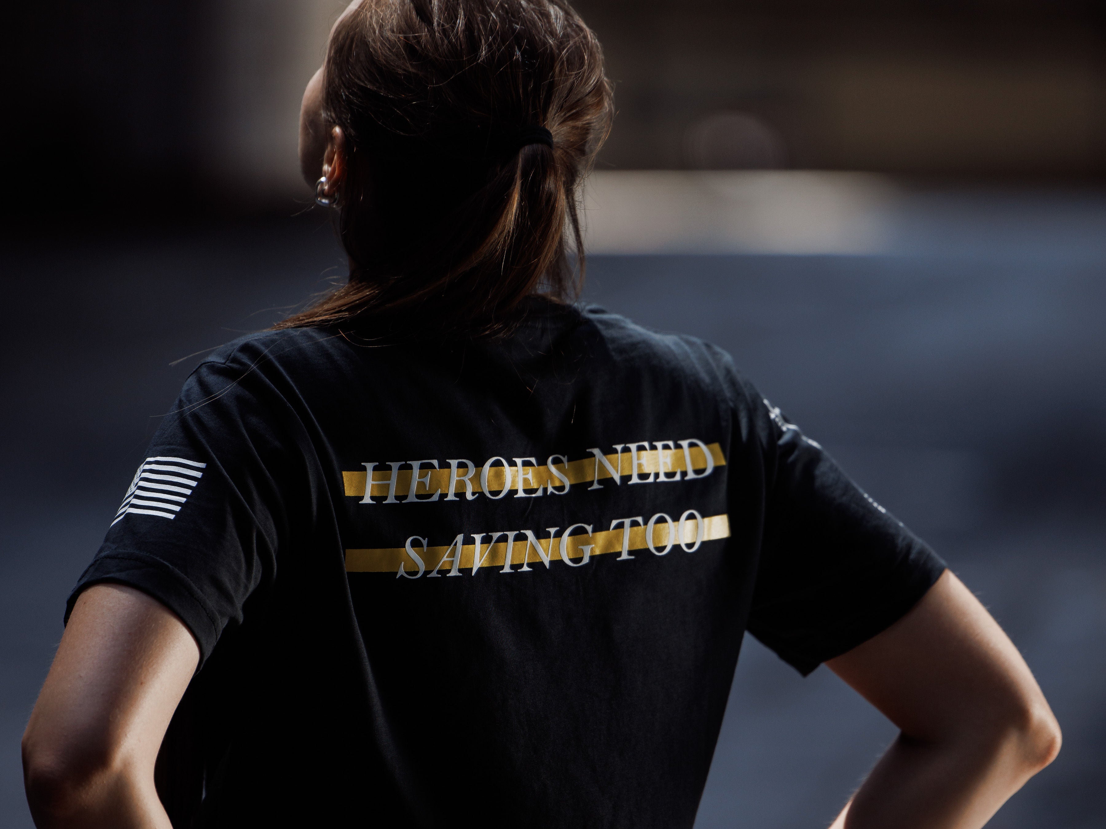 Person wearing a black t-shirt with 'HEROES NEED SAVING TOO' text on the back, standing against a dark background.