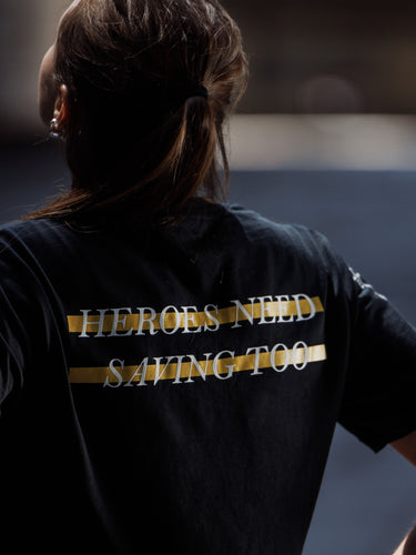 Person wearing a black t-shirt with 'HEROES NEED SAVING TOO' text on the back, standing against a dark background.